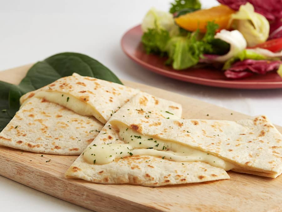 A wooden cutting board with a folded flatbread and a plate of mixed greens and vegetables in the background.