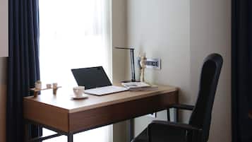 Une chambre d’hôtel avec un bureau en bois, une chaise de bureau noire, un ordinateur portable, une petite plante en pot, une tasse et une poubelle.