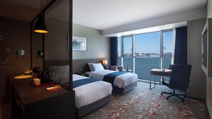 A hotel room with two beds, a desk, a chair, a large window with a view of the water, and a bathroom with a sink and mirror. - Asti Hotel Busan Station (Busan)