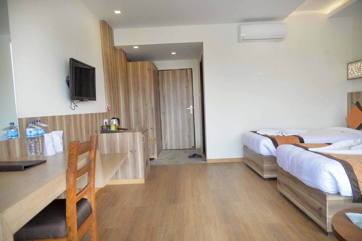 A hotel room with a wooden desk, two beds, a TV, and a shower area.