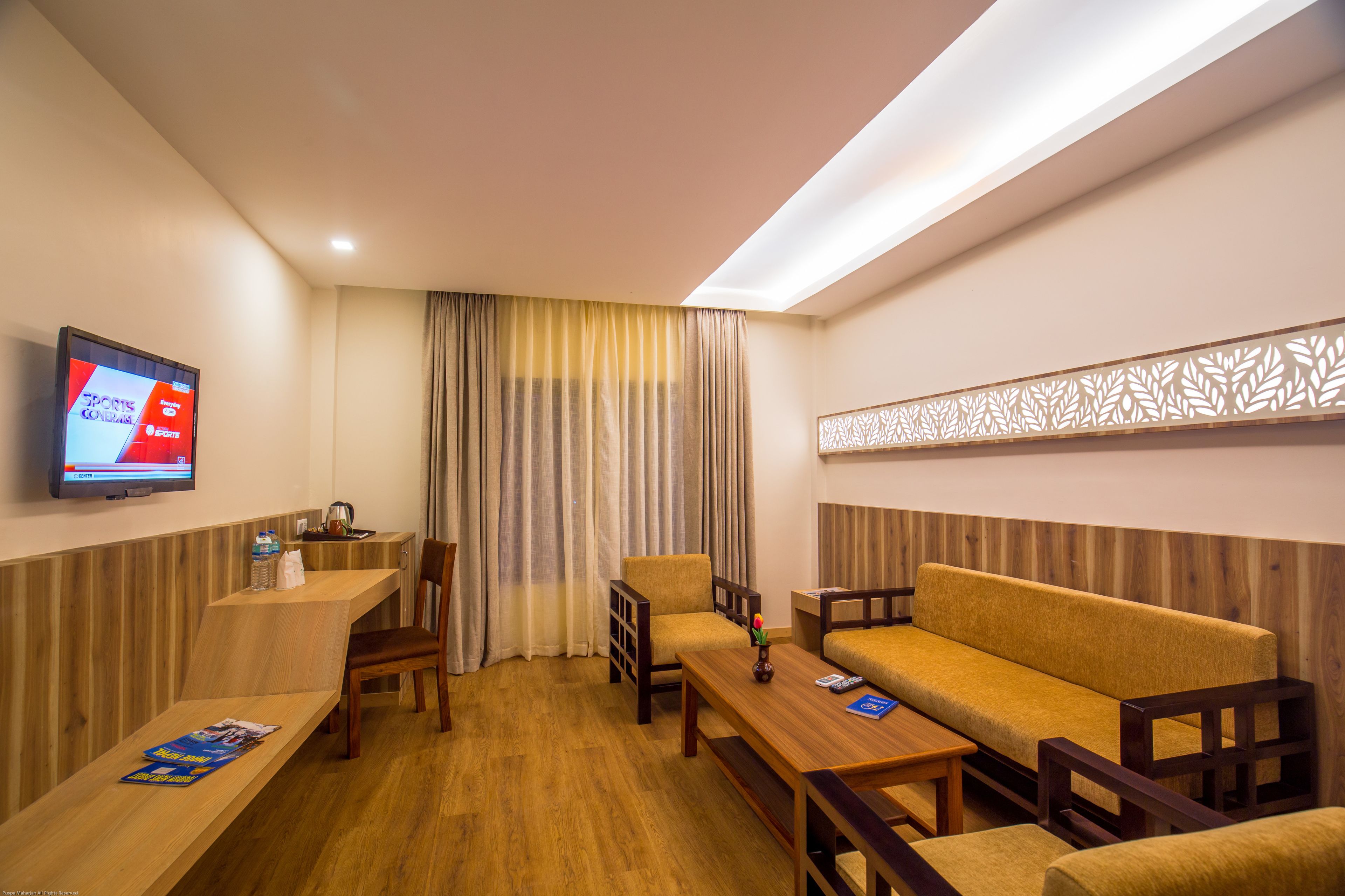 A hotel room with a wooden floor, a sofa, a chair, a small table, a TV mounted on the wall, and a curtain.