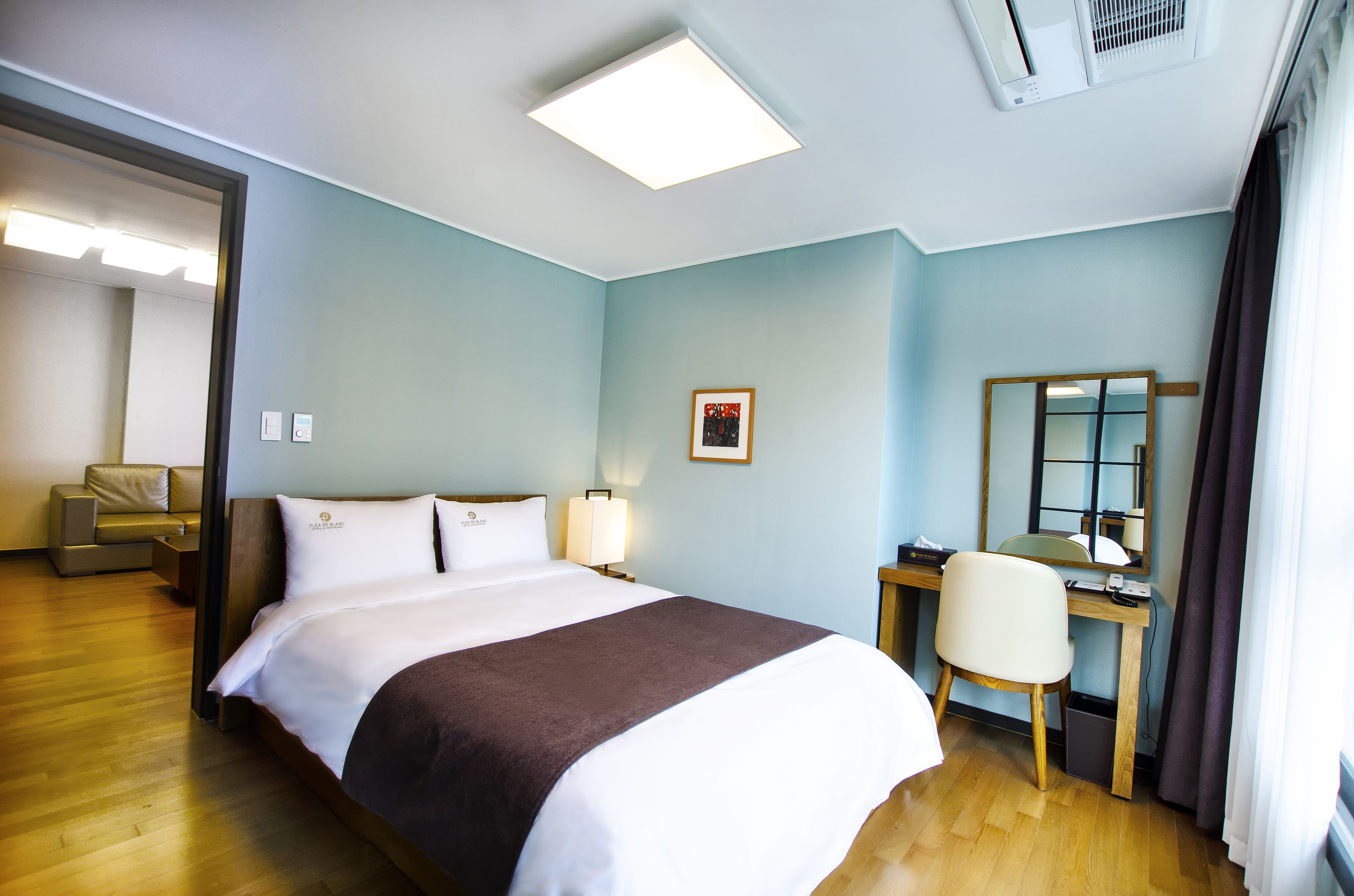 A hotel room with a bed, a desk, a chair, a desk lamp, a framed picture, and a window with curtains.