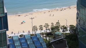 A beach with people, a building with a glass facade, and a clear sky. - Plea De Blanc (Busan)