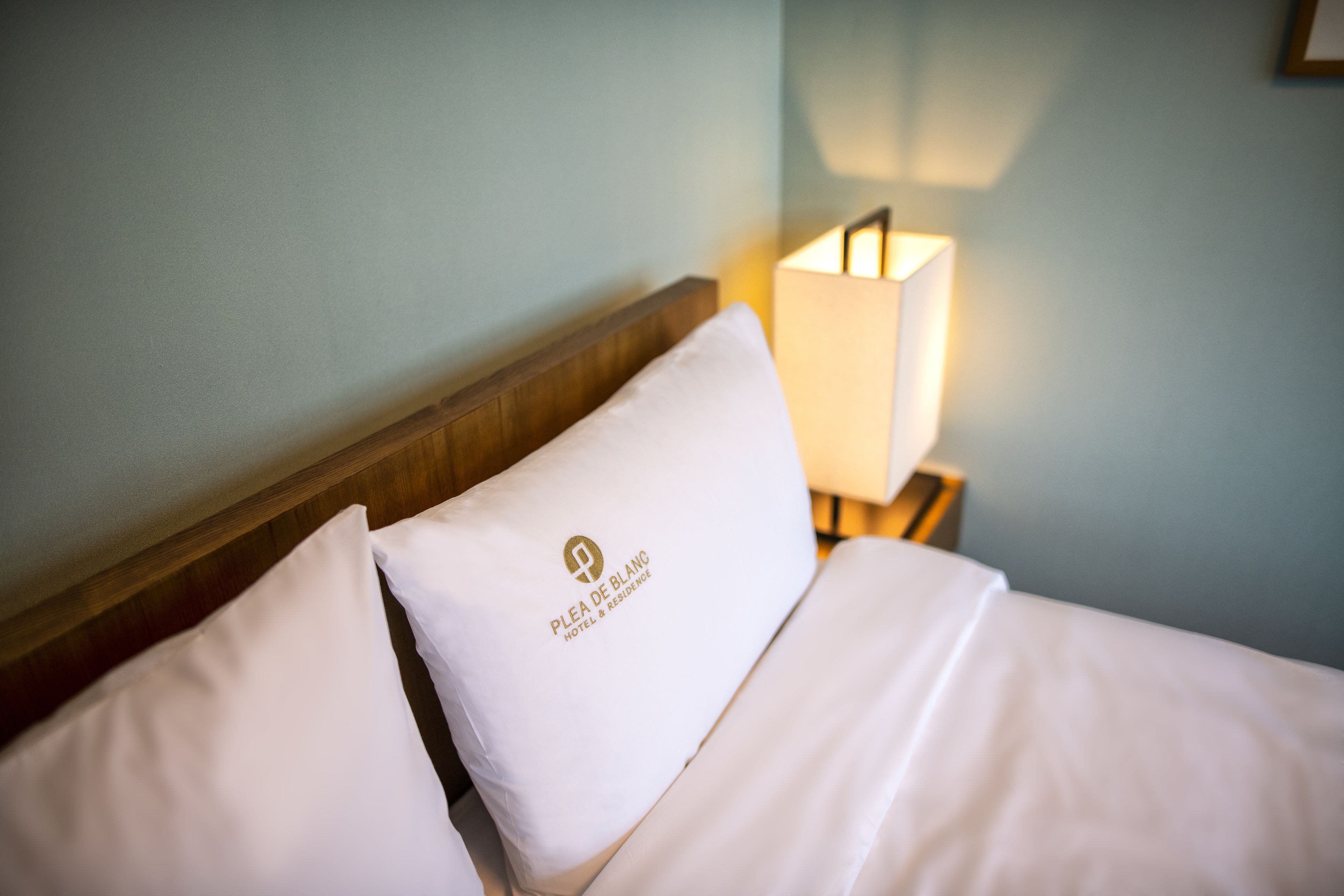 A neatly made bed with white linens and a pillow with a logo, a bedside lamp, and a wooden headboard.