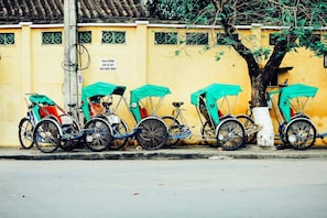 A row of rickshaws parked against a yellow wall with decorative tiles and a tree. - Hoi An Green Town House Homestay (Da Nang)