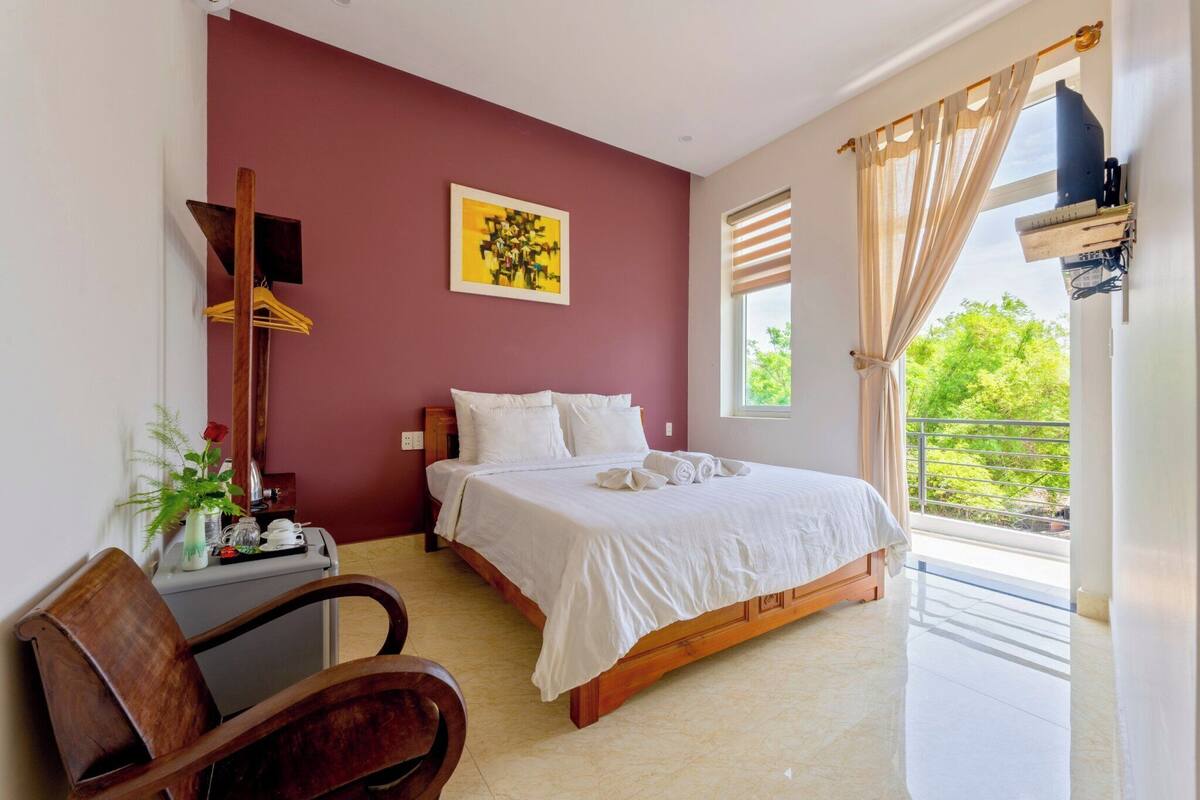 A bedroom with a bed, a desk, a chair, a window with curtains, and a painting on the wall.