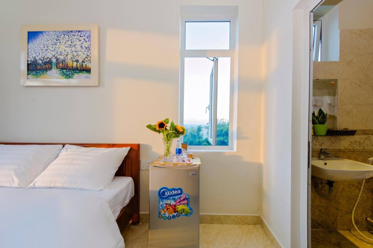 A hotel room with a bed, a small table with a vase of flowers, a window, and a bathroom with a sink and mirror.