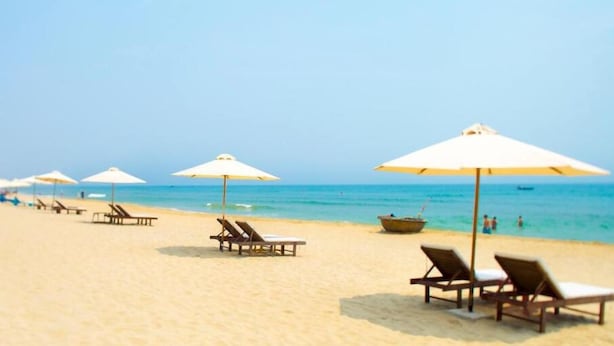 A beach with rows of sun loungers and umbrellas, a boat on the water, and a clear sky. - Hoi An Green Town House Homestay (Da Nang)