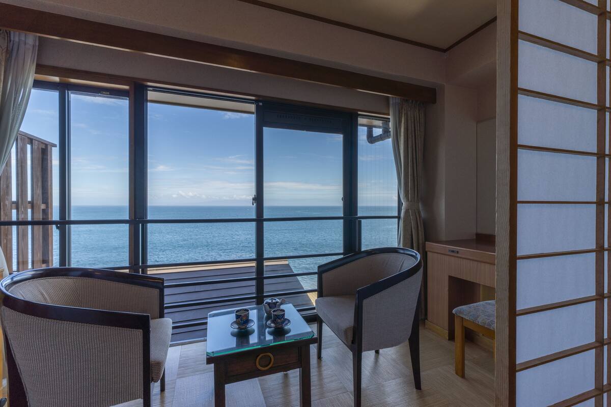 A balcony with a view of the sea, two chairs, a table with a teapot and cups, and a wooden desk.