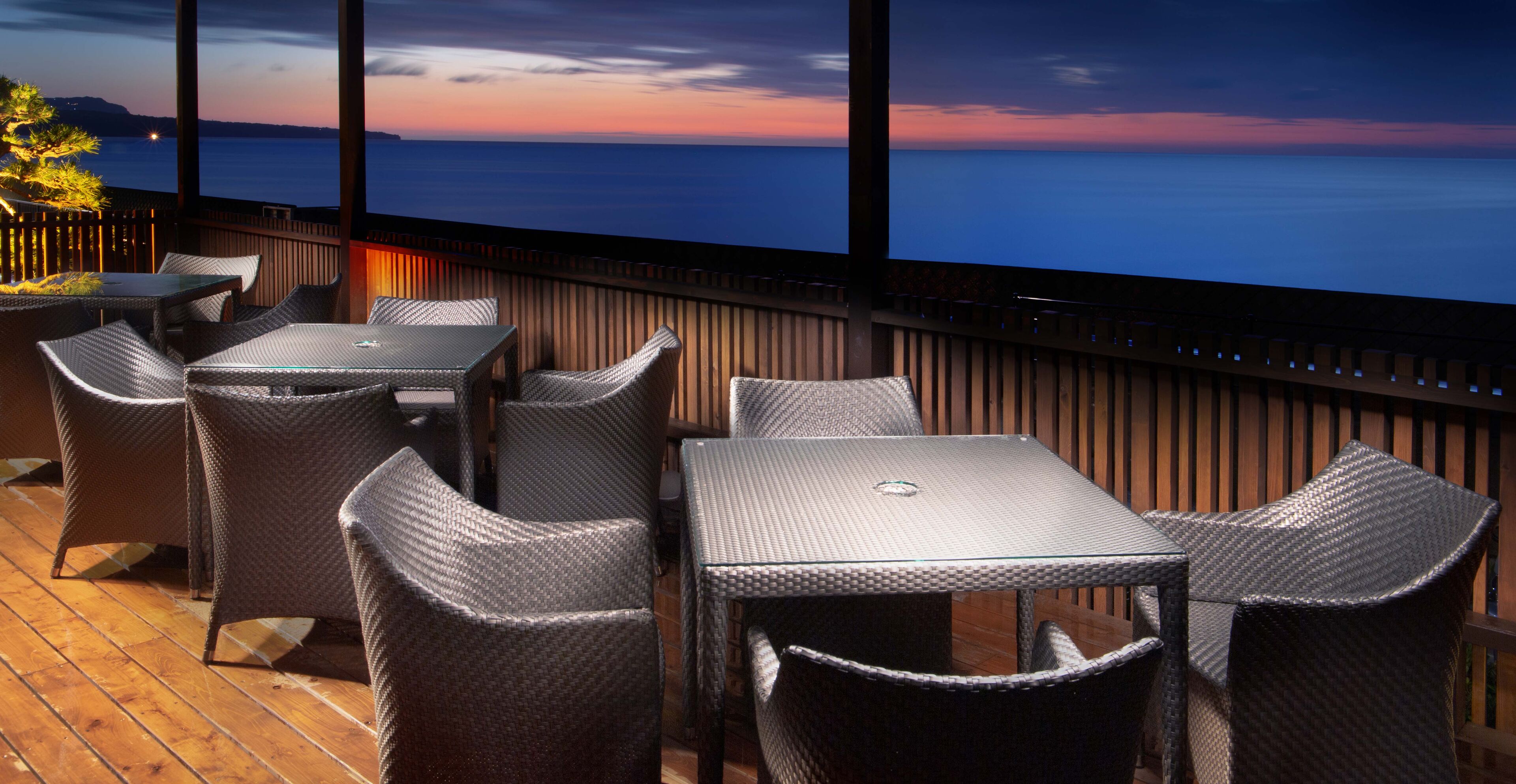 A terrace with wicker furniture and a glass table, overlooking a body of water at sunset.