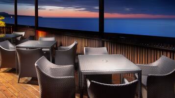 A terrace with wicker furniture and a glass table, overlooking a body of water at sunset.