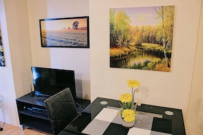 A room with a flat-screen TV on a stand, a glass table with a checkered table runner, and a vase with yellow flowers.