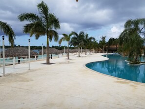 Área de piscina en un resort, con palmeras, una estructura de techo de paja y vista despejada del océano.