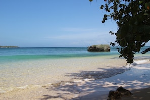 Ein Strand mit klarem türkisblauem Wasser, einem Sandufer und einer großen Felsformation im Hintergrund.