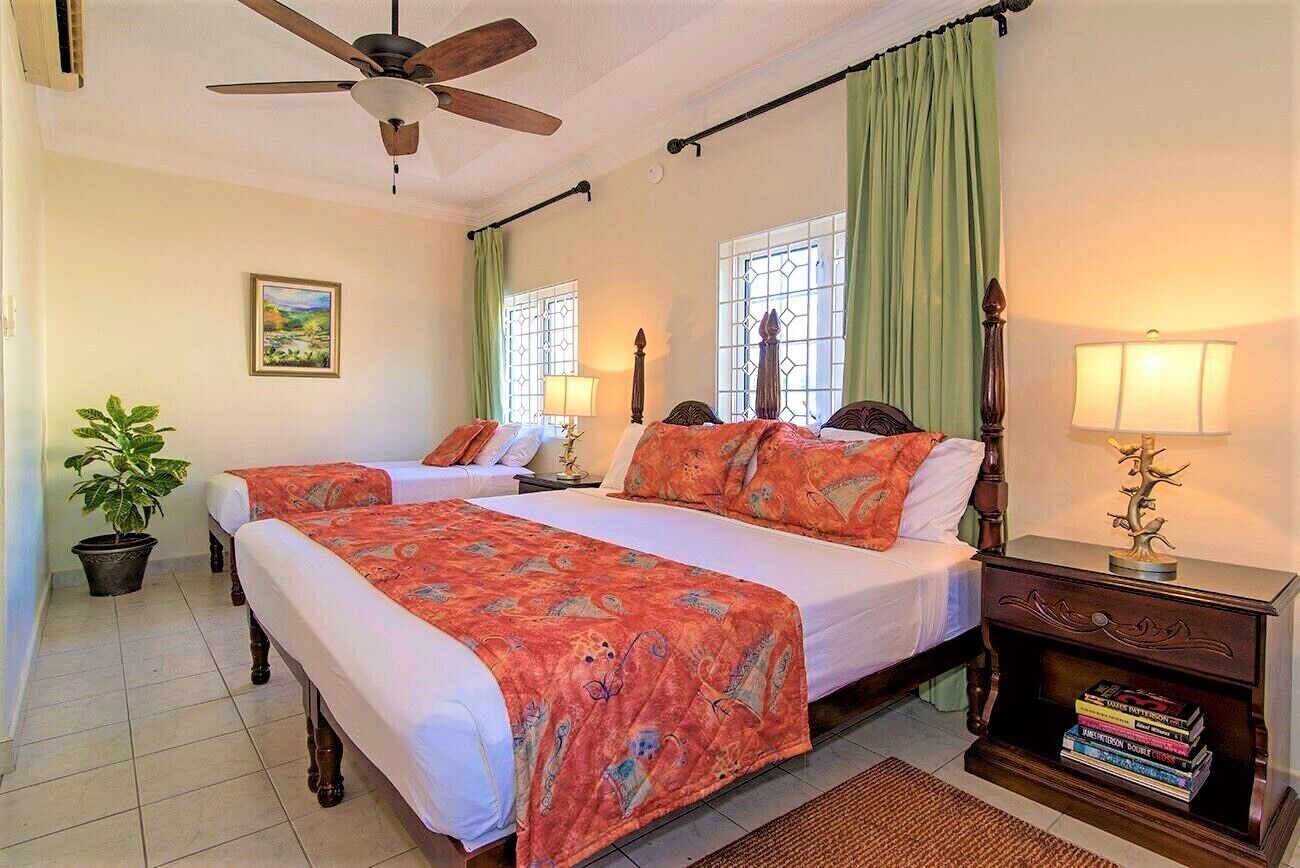 A hotel room with two beds, a ceiling fan, a potted plant, and a nightstand with books.