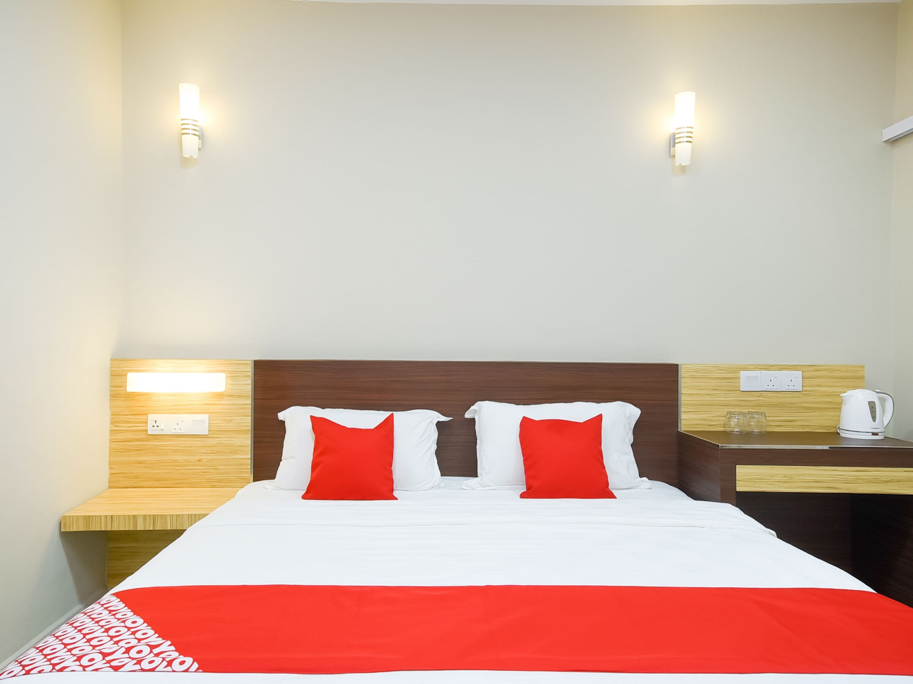 Una habitación de hotel con cabecera de madera, dos mesitas de noche, una cama blanca con almohadas rojas y una manta roja.