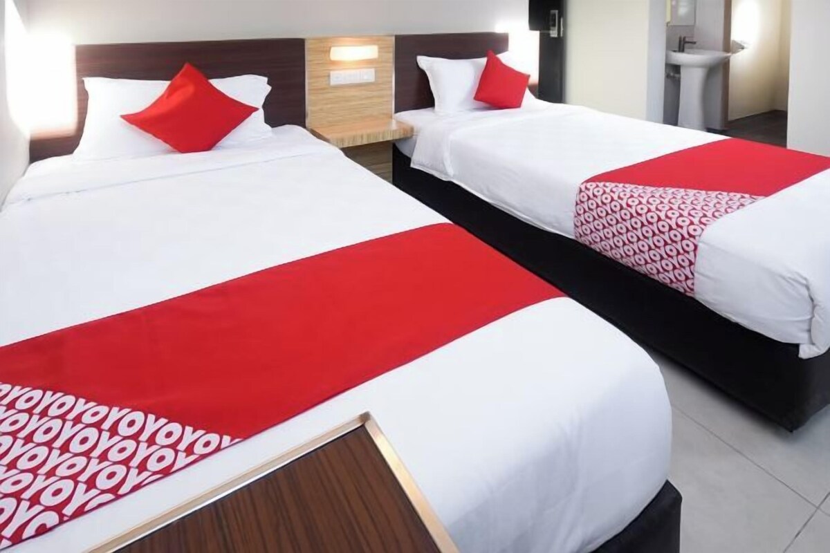 Una habitación de hotel con dos camas, cada una con ropa de cama blanca y detalles en rojo. Hay un cabecero de madera, una mesita de noche y una lámpara de pared.