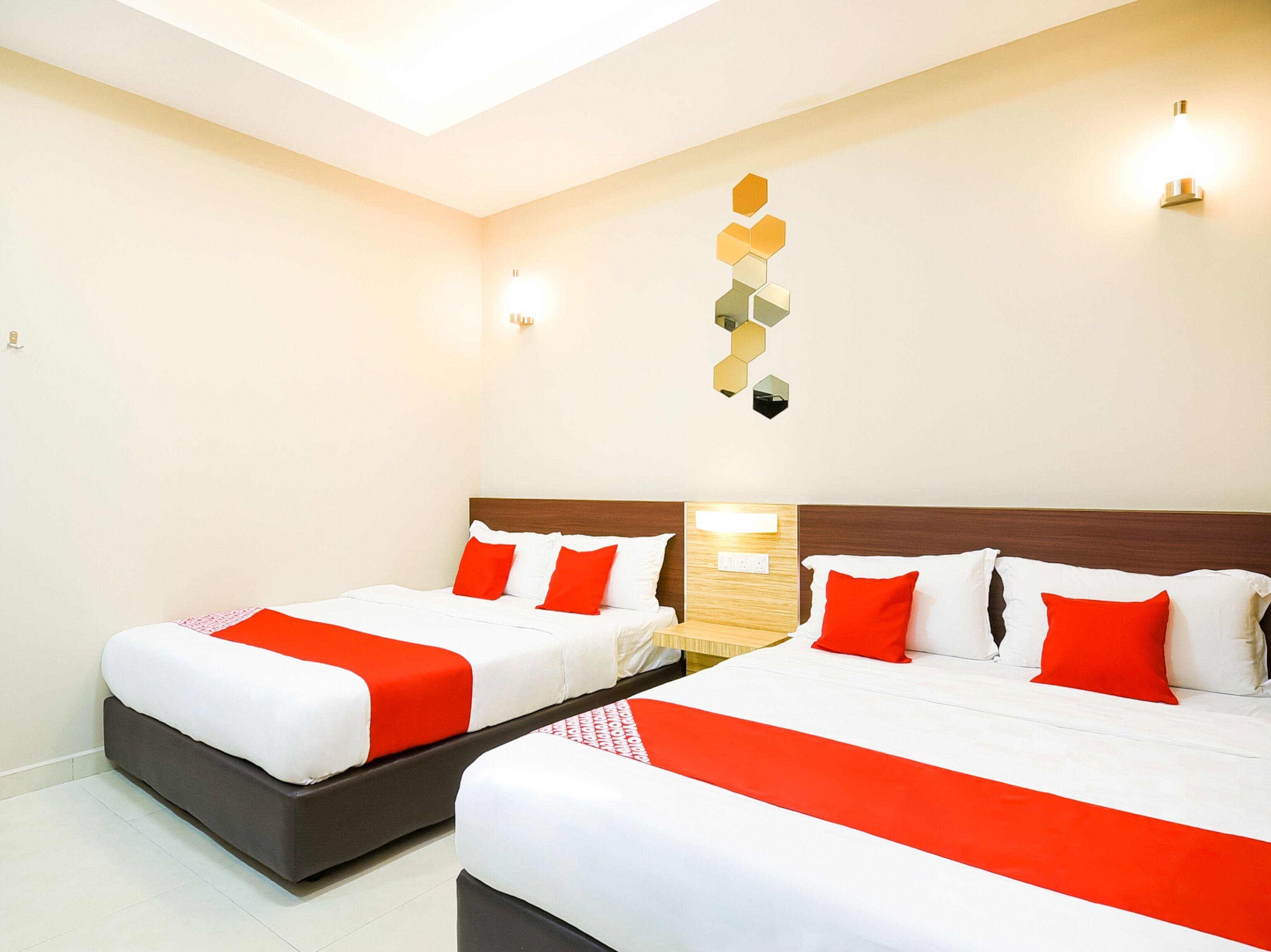 A hotel room with two beds, each with white bedding and red accents, a wooden headboard, and a wall-mounted light fixture.