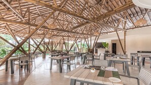 A spacious indoor dining area with bamboo ceiling beams, multiple tables and chairs, and a view of greenery outside. - Daun Lebar Villas (Payangan)