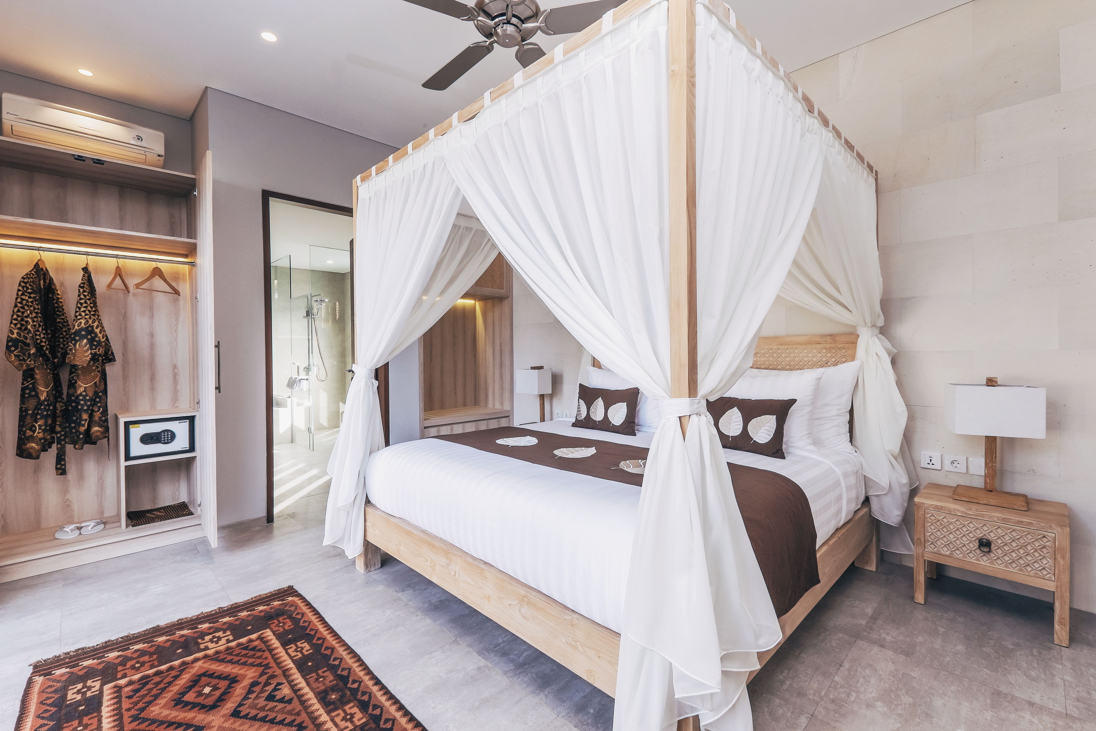 A four-poster canopy bed with a wooden frame and white drapes, a bedside table with a lamp, and a patterned rug on the floor.
