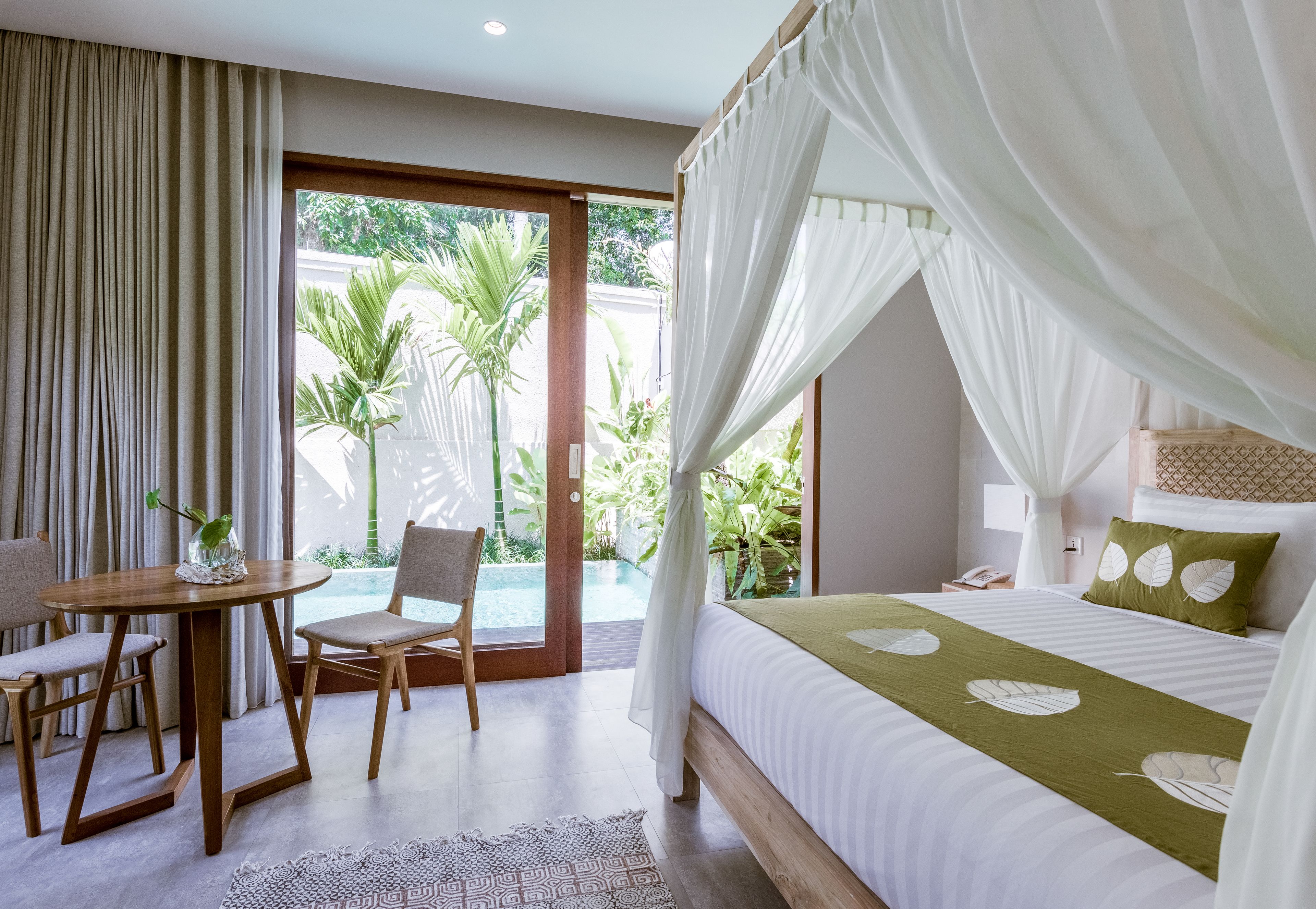Une chambre avec un lit, une table de chevet, une chaise, une petite table et une vue sur une piscine et la verdure environnante.