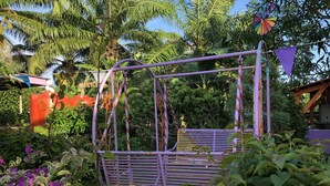 Een paarse schommel in een weelderige tuin met diverse planten en een palmboom op de achtergrond.