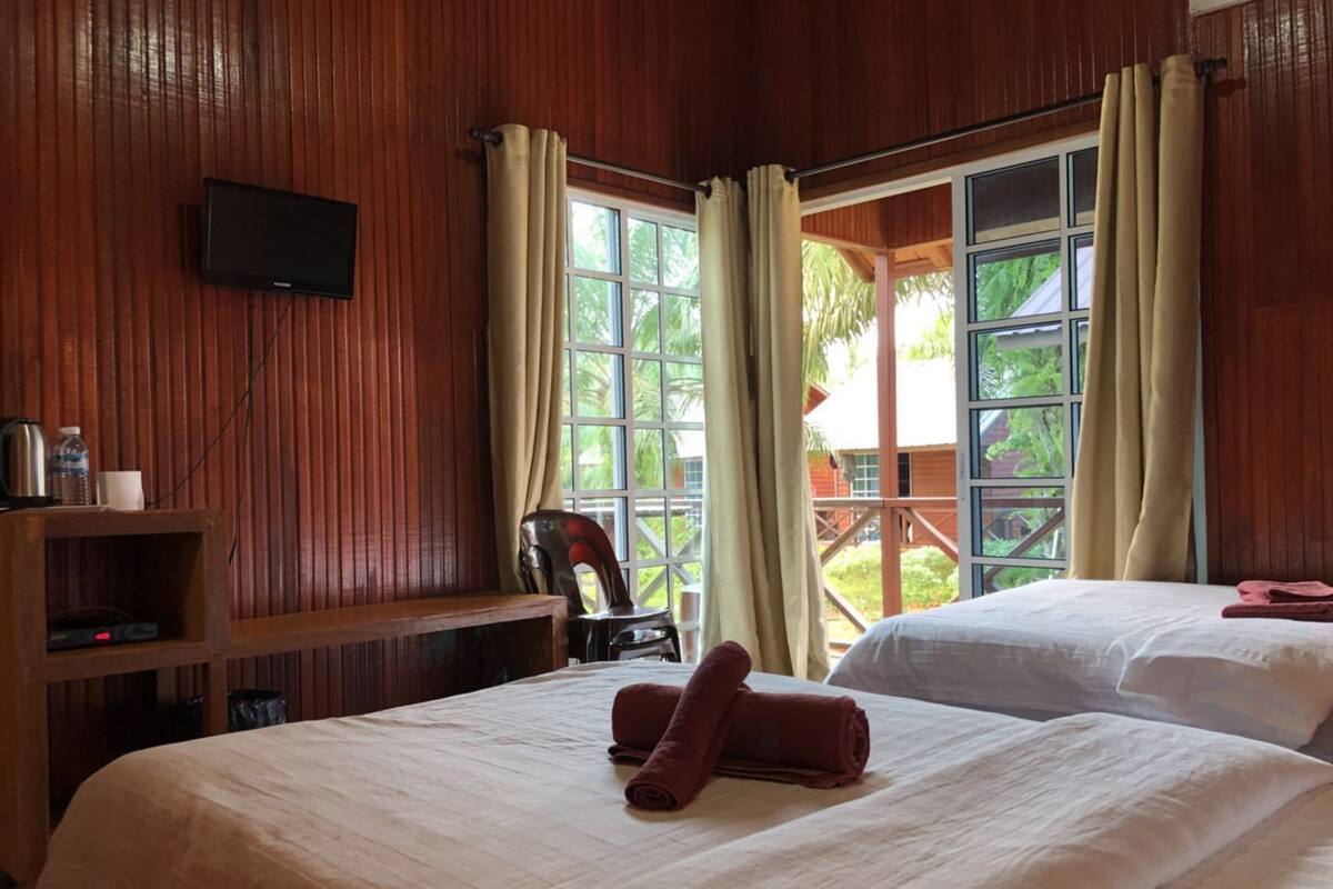 Habitación de hotel con dos camas, paredes de madera, televisor montado y balcón con cortinas.