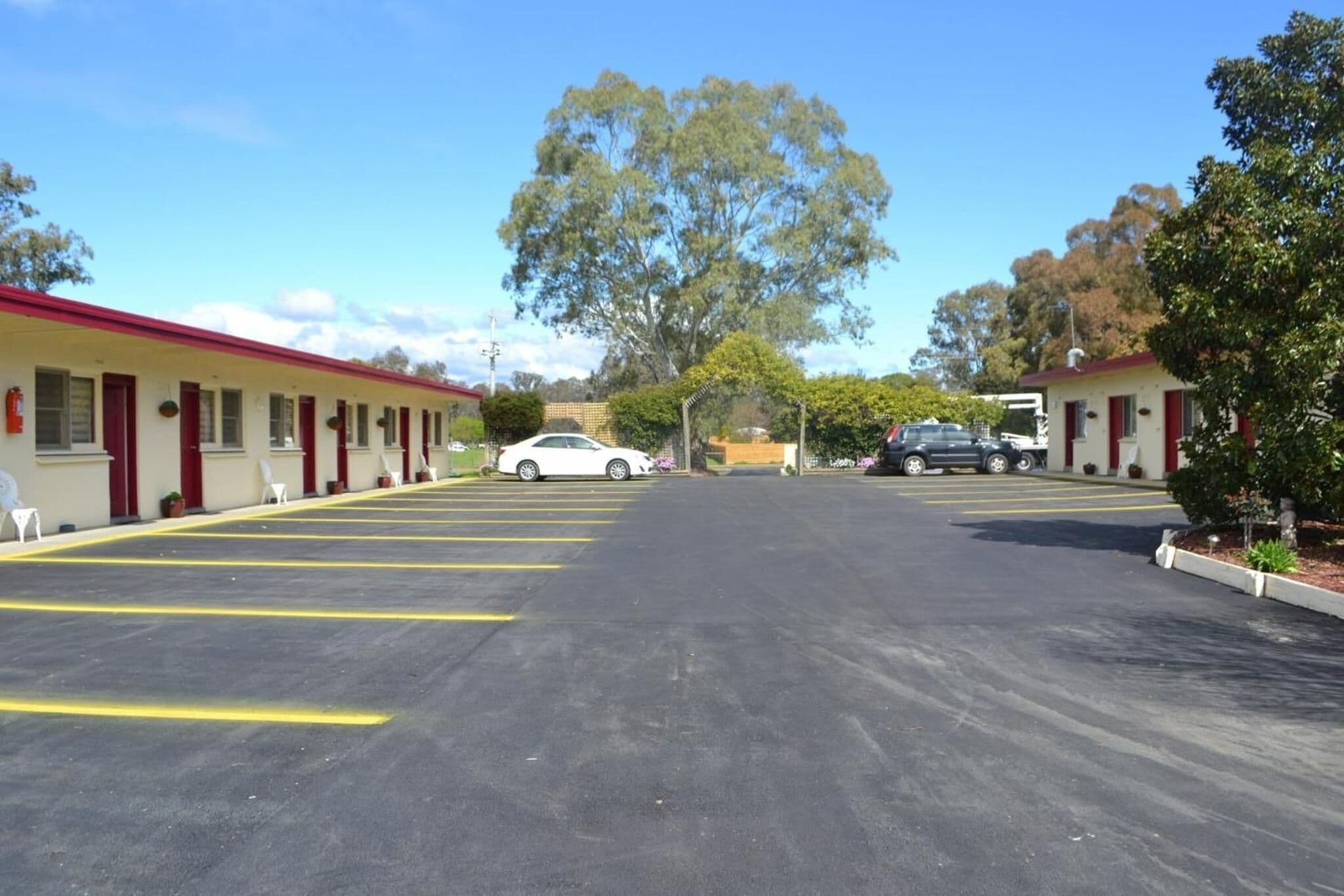 A motel with a row of single-story buildings, each with a red roof and white walls. There are several cars parked in the lot, and a large tree is visible in the background.