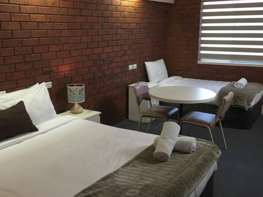 A hotel room with a brick wall, a bed with white linens, a round table with chairs, and a window with blinds.