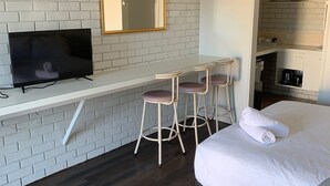 A modern hotel room with a white brick wall, a flat-screen TV, a small bar area with stools, a bed with white linens, and a visible kitchen area. - Wattle Motel (Seymour)