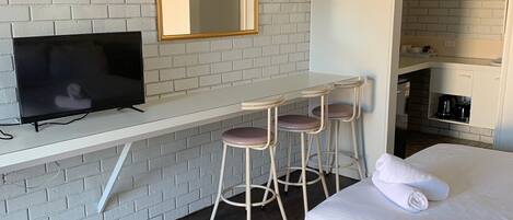 Una habitación de hotel moderna con pared de ladrillo blanco, televisor de pantalla plana, un pequeño bar con taburetes, cama con ropa de cama blanca y una zona de cocina visible.