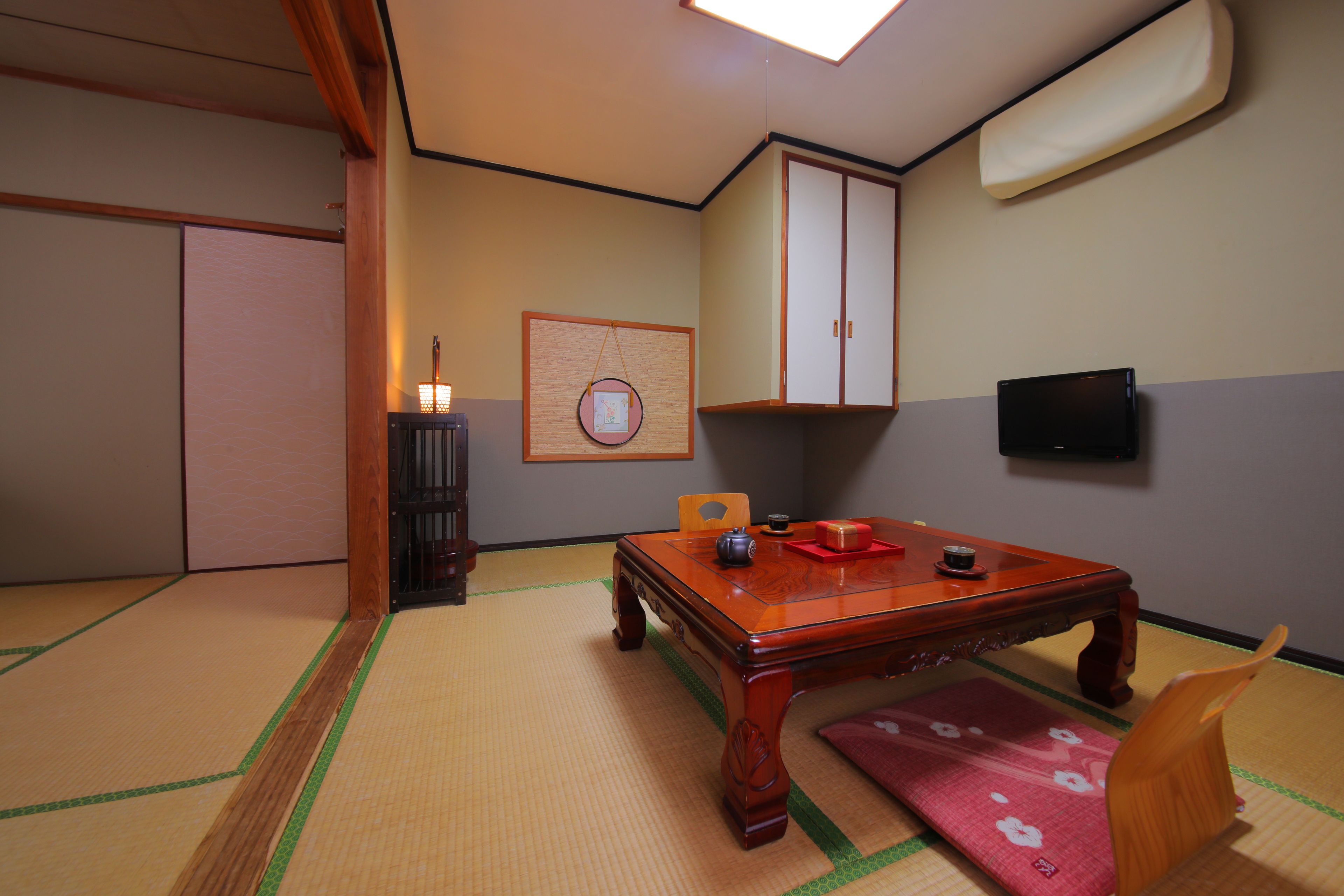 Una habitación japonesa tradicional con suelo de tatami, una mesa de madera baja, un cojín rojo y un televisor montado en la pared.