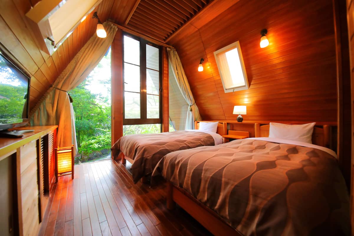 Quarto de cabana de madeira com duas camas, uma escrivaninha pequena e uma janela com vista para o verde.