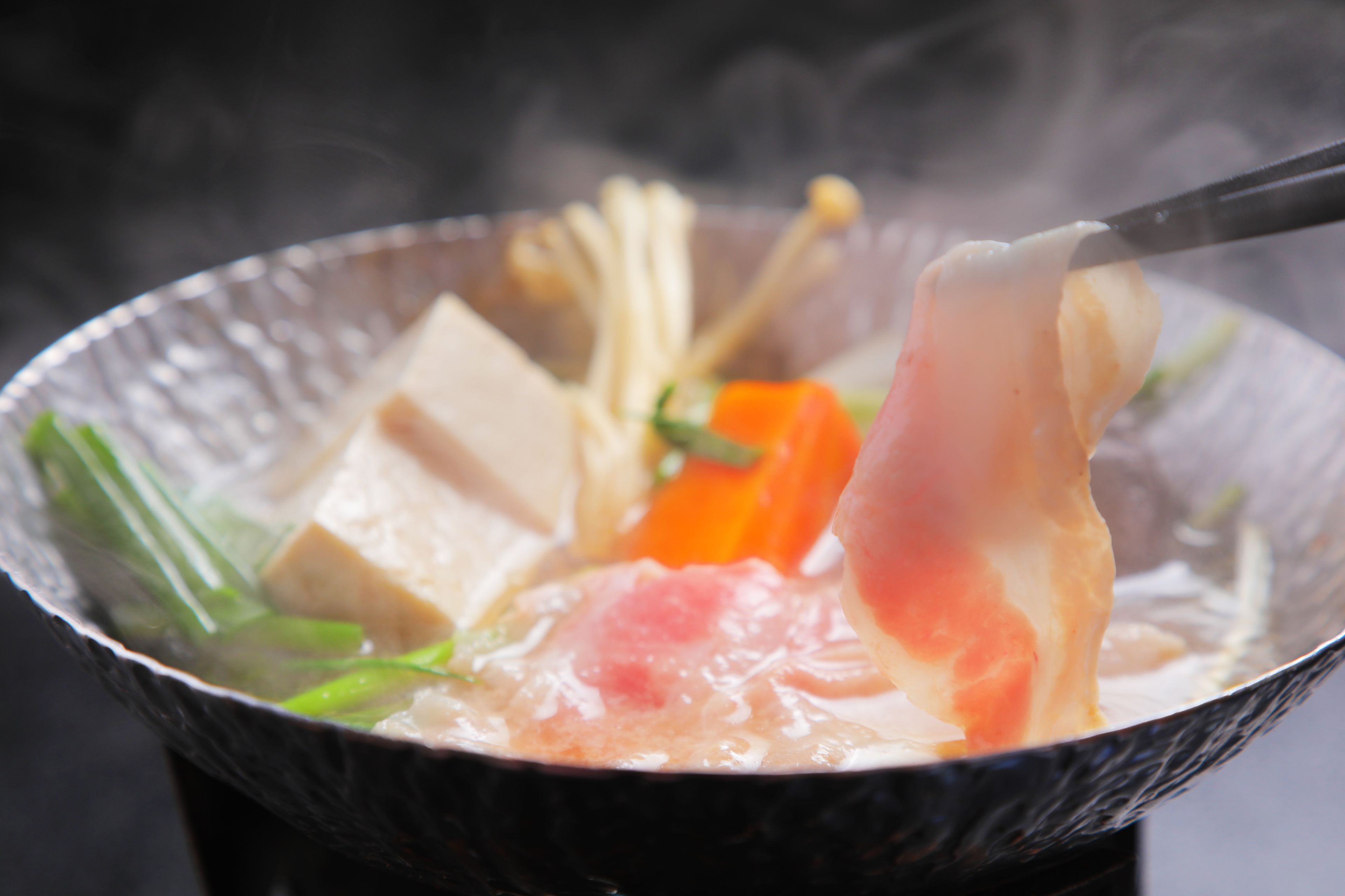 A close-up of a hot pot meal with tofu, vegetables, and meat being lifted with chopsticks.