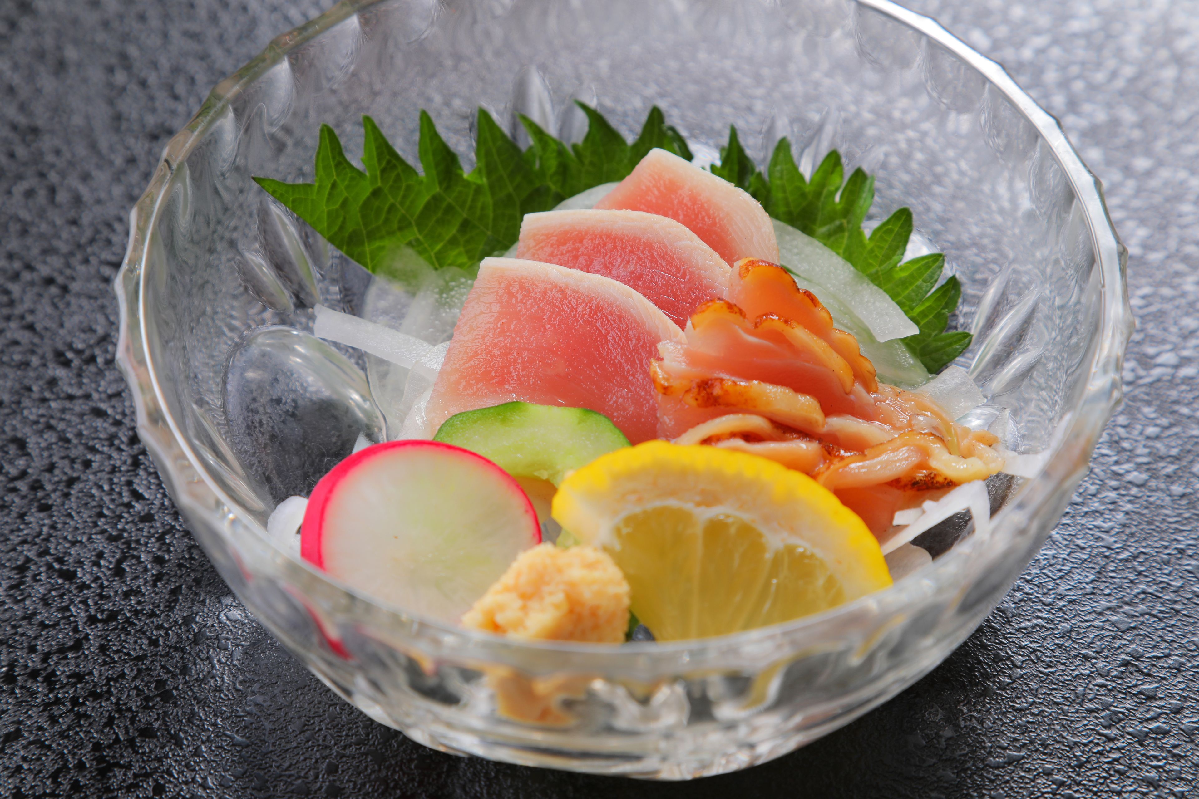 A clear glass bowl containing slices of pink fish, green leaves, radish, lemon, and a piece of orange-colored food on a textured surface.