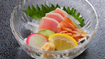 A clear glass bowl containing slices of pink fish, green leaves, radish, lemon, and a piece of orange-colored food on a textured surface.