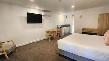 Habitación de hotel con cama, televisor montado en la pared, kitchenette pequeña y zona de estar con una silla y mesa.