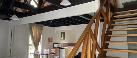 A room with a bed, a dining table with chairs, a kitchen area, and a wooden staircase.
