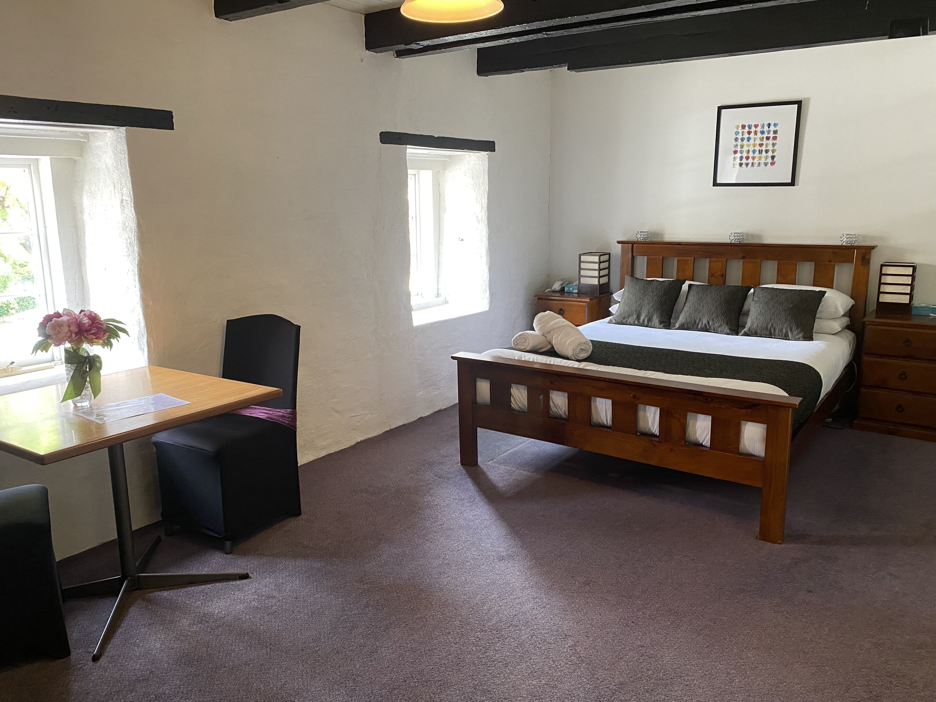 Une chambre à coucher avec un lit en bois, des tables de chevet, un bureau avec une chaise et une fenêtre agrémentée d’un vase de fleurs.
