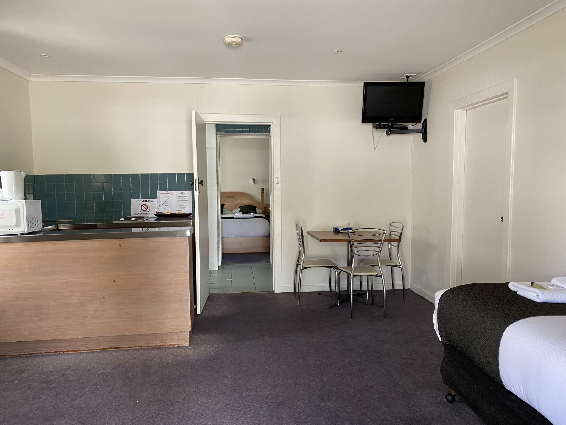 Una habitación de hotel con kitchenette, una cama, una mesa de comedor con sillas y un televisor montado en la pared.