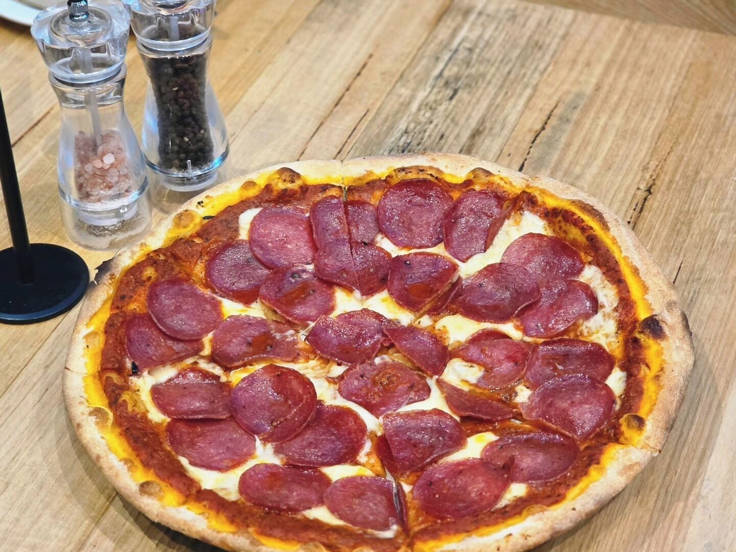 Una pizza de pepperoni sobre una mesa de madera, con saleros y pimenteros cerca.