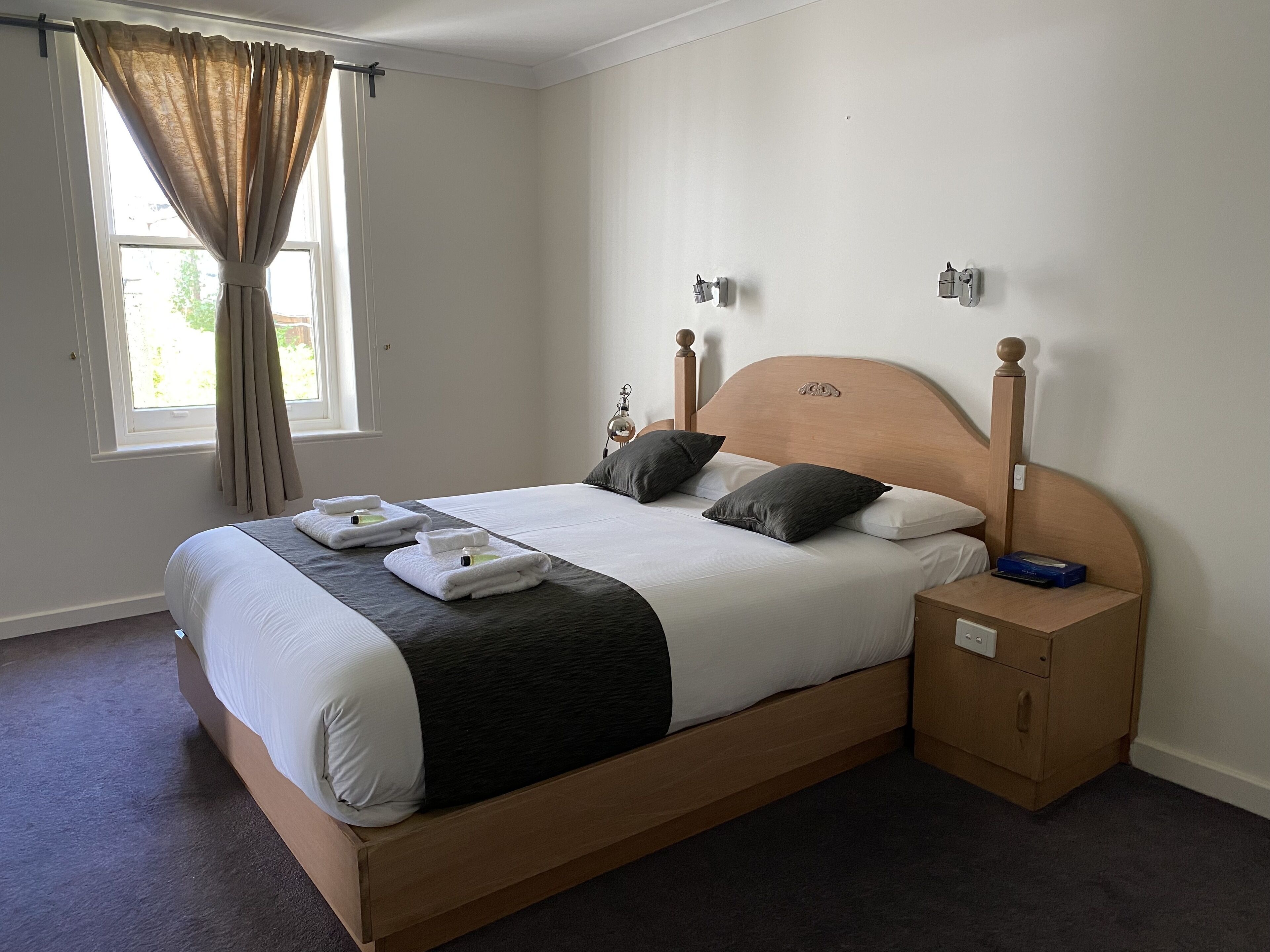 A neatly made bed with a wooden headboard, a bedside table with a lamp, and a window with curtains.