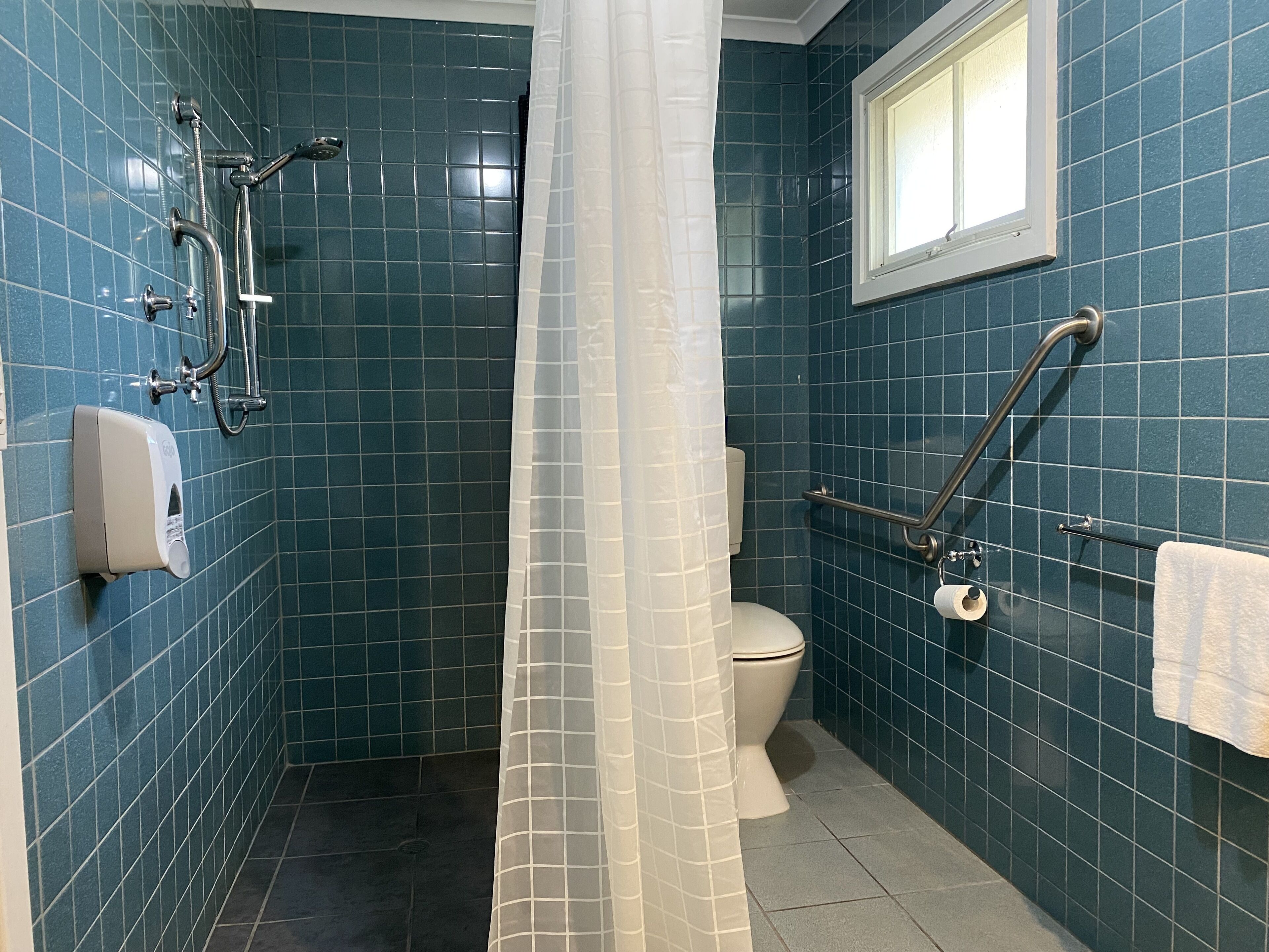 A bathroom with blue tiles, a toilet, a hand dryer, a shower with a glass door, and a window.