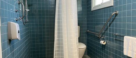 A bathroom with blue tiles, a toilet, a hand dryer, a shower with a glass door, and a window.