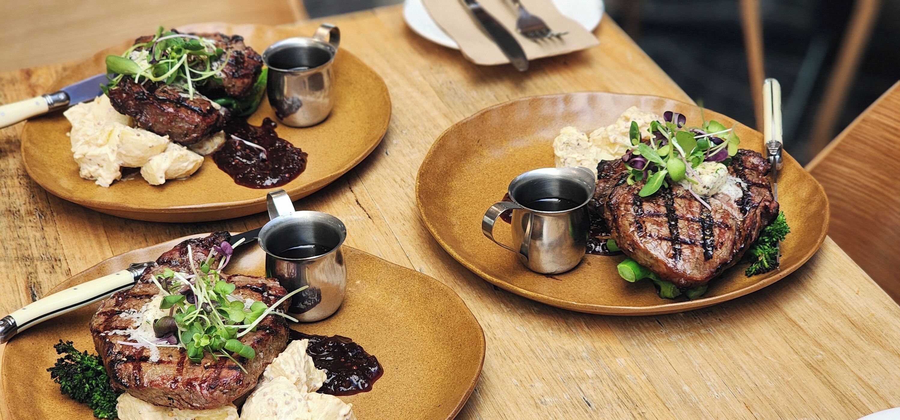 A wooden table with three plates of steak, each with a small metal cup and a knife.