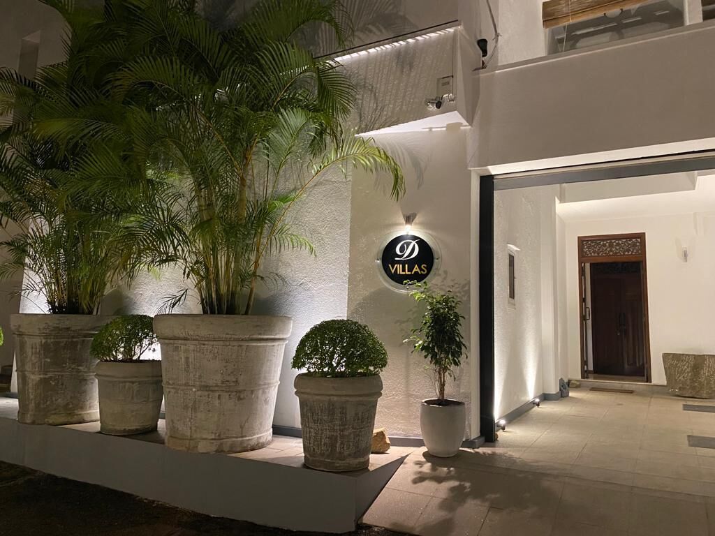 Un ingresso ben illuminato con un cartello che riporta la scritta "VILLAS" e grandi piante in vaso che fiancheggiano il vialetto.