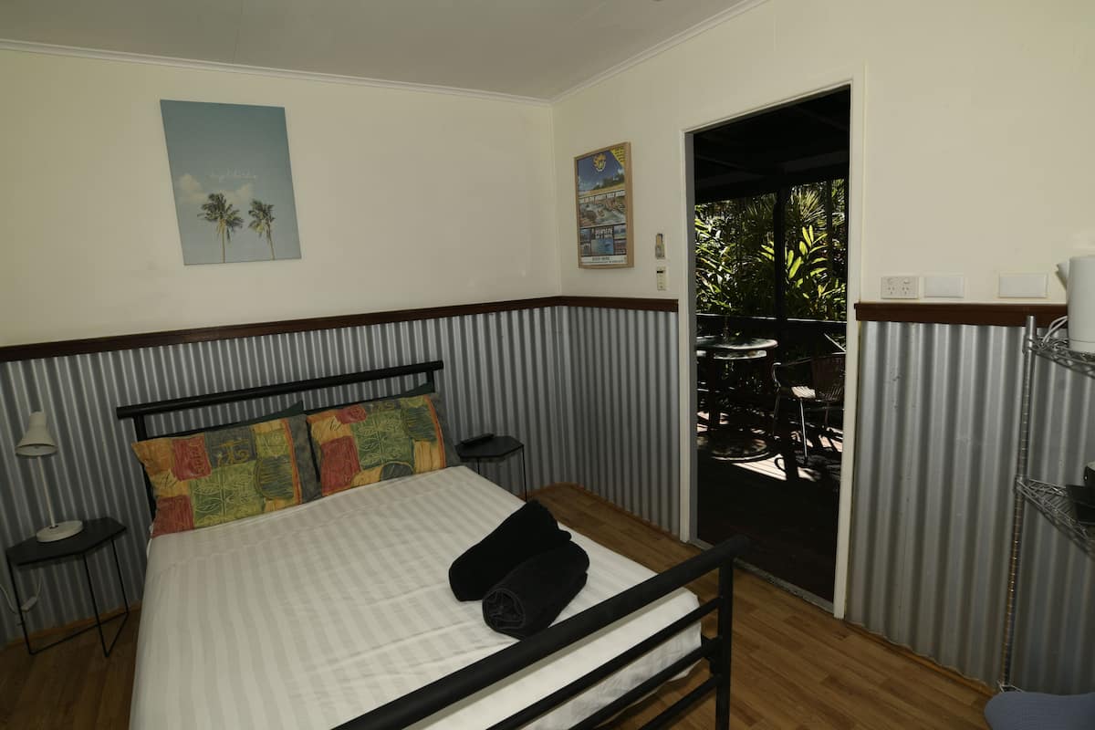 A bedroom with a bed, a nightstand, a lamp, a window with a view of palm trees, and a door leading to an outdoor area.