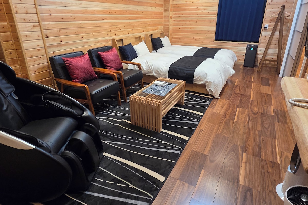 Una habitación tipo cabaña de madera con una cama, un sillón de masaje, una mesita y una ventana con cortinas.