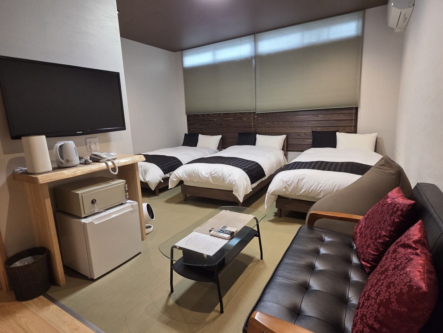 Quarto de hotel com duas camas, televisão, secretária com telefone, mini-frigorífico, máquina de café e sofá de couro.