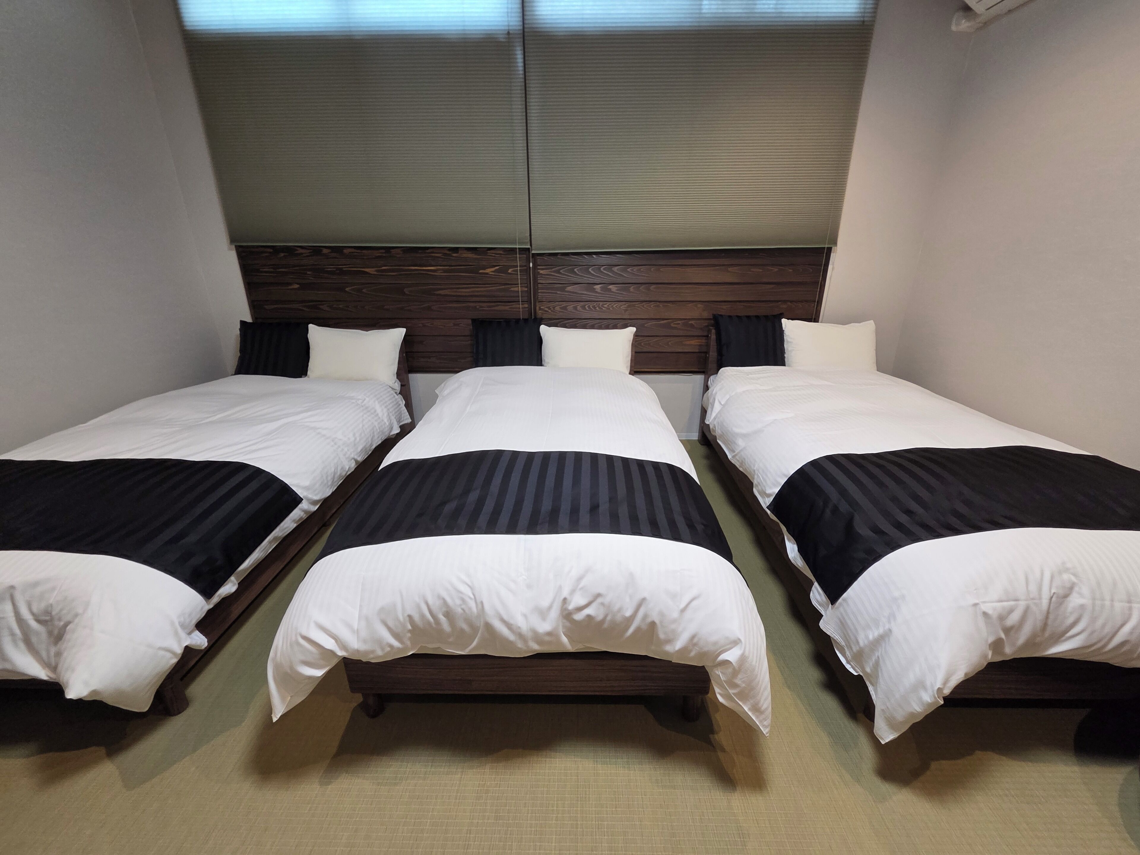A hotel room with three single beds, wooden headboards, and a large window with blinds.